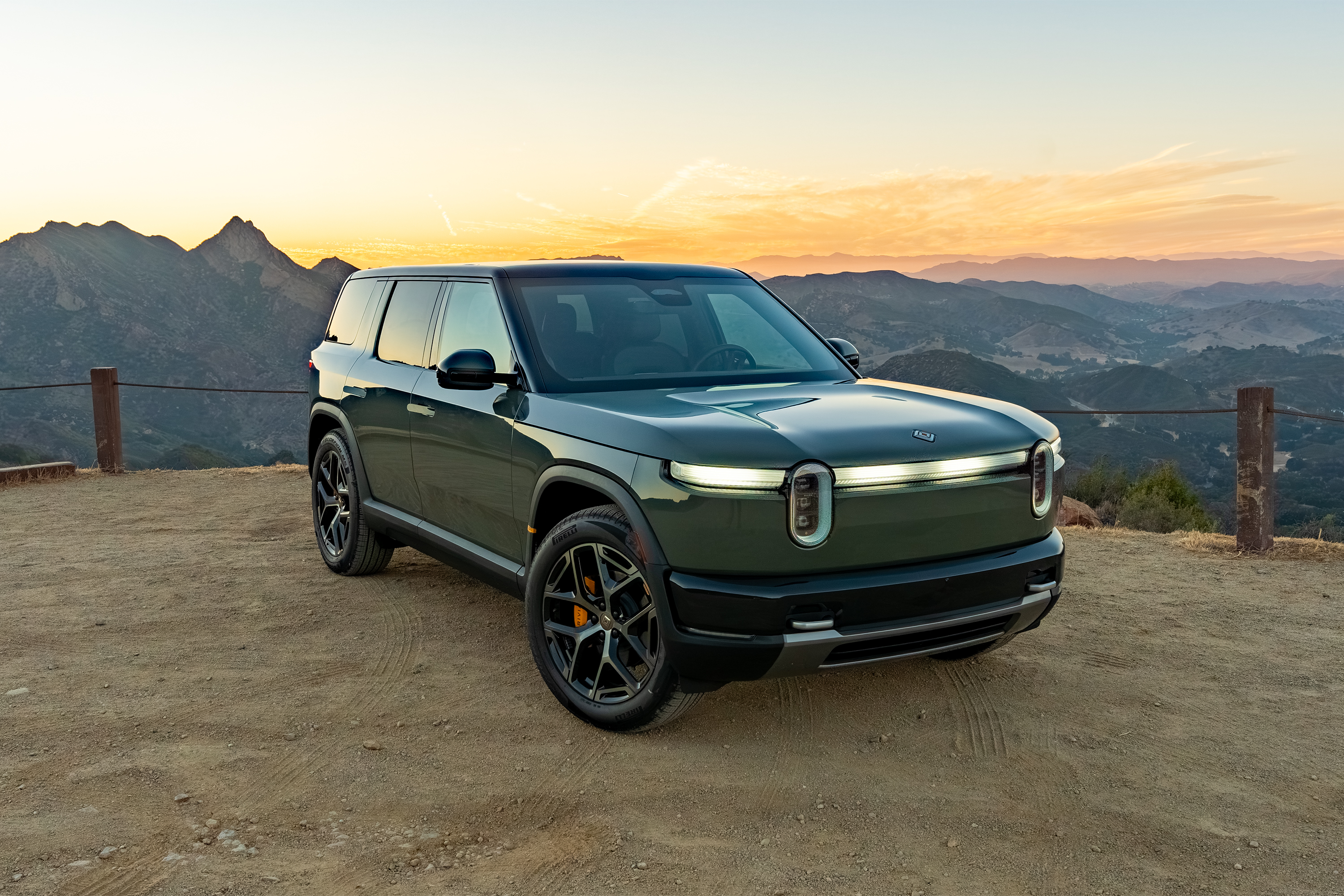 Rivian R1S electric SUV parked on a mountain overlook at sunset with rolling hills in background