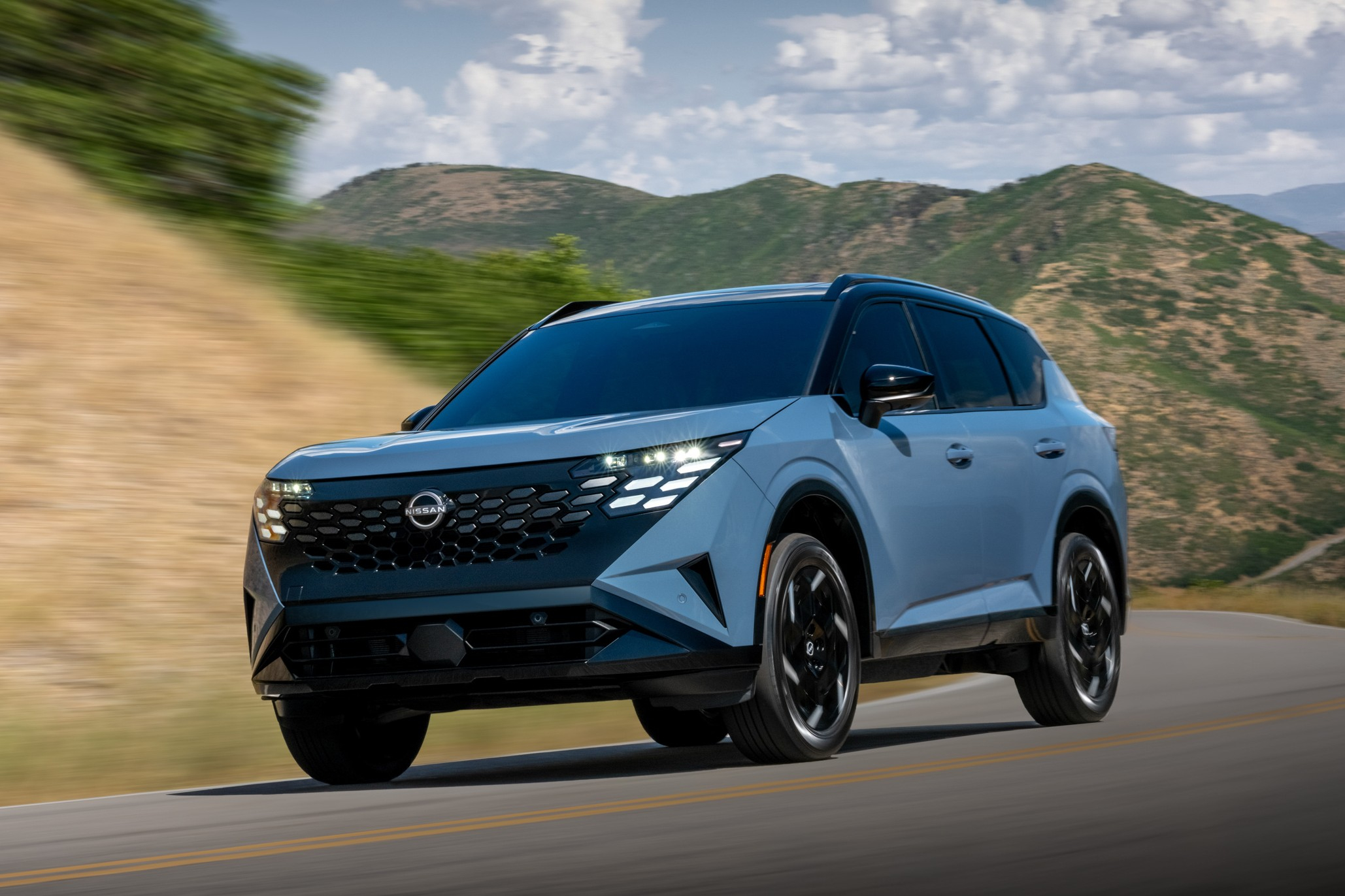 Light blue Nissan Ariya electric SUV driving on a mountain road with scenic hills in the background