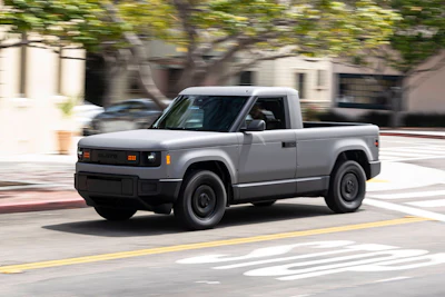 Silver Rivian R1T electric pickup truck driving on urban street with trees and buildings