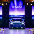 Luxury sports car displayed on stage during brand launch event with illuminated screens and dramatic blue lighting