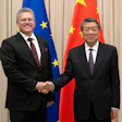 Two officials in formal business attire shake hands at a diplomatic meeting with EU and Chinese flags in the background.