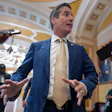 Man in blue suit and gold tie speaking to media in ornate government building corridor