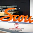 Scout Motors orange brand signage displayed outside showroom with tan SUV and Manufacturing Now logo