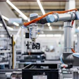 Two orange and silver robotic arms working together in a modern manufacturing facility with white structural elements and industrial equipment.