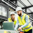 Two construction workers in hard hats and yellow safety vests review plans on a laptop inside an industrial warehouse facility.