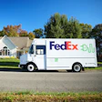 FedEx delivery truck parked on residential street in front of white house with autumn trees