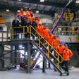 Industrial facility tour with group in orange safety uniforms on yellow metal stairs and platforms