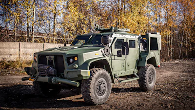 Tactical armored military vehicle with open door parked outdoors against autumn trees