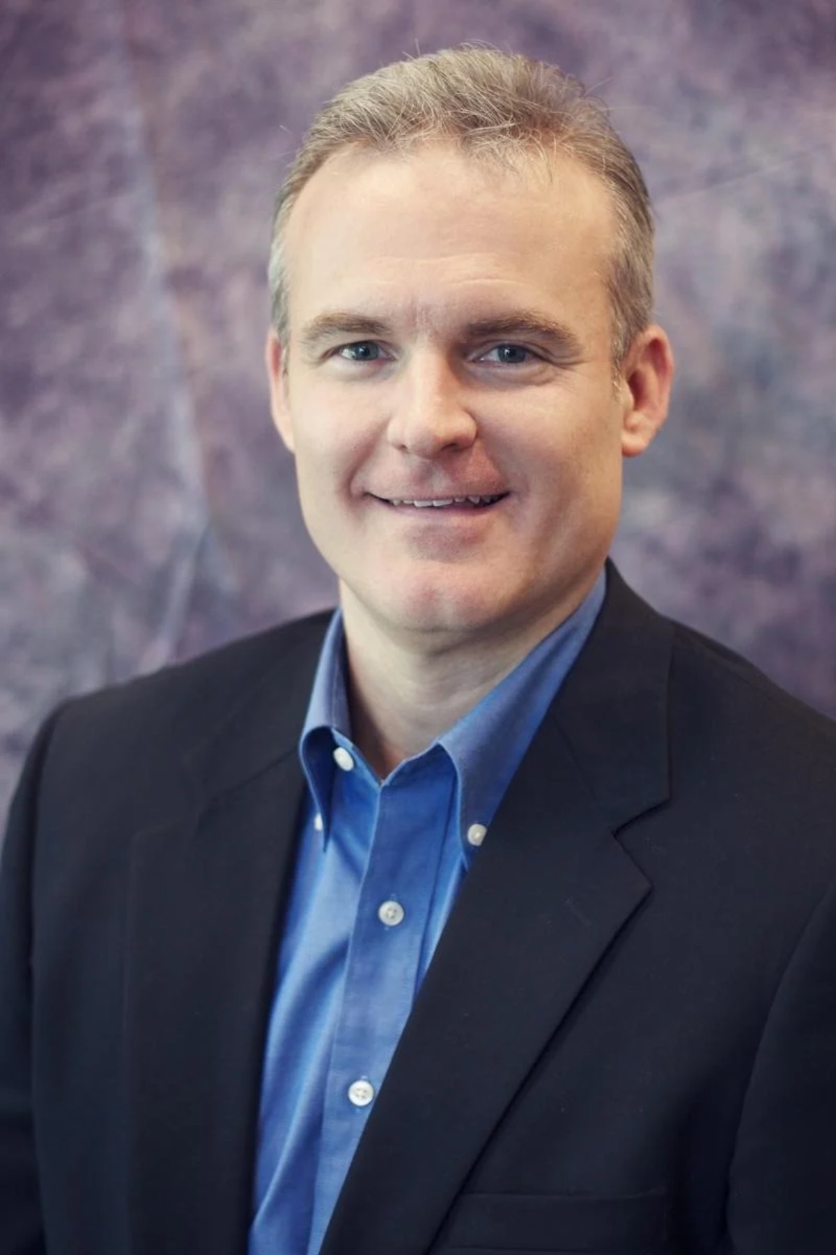 Professional headshot of a man in a dark blazer and blue button-up shirt smiling at the camera Headshot