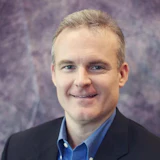 Professional headshot of a man in a dark blazer and blue button-up shirt smiling at the camera