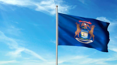 Michigan state flag waving on a flagpole against a blue sky with white clouds