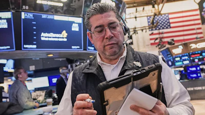 Trader at NYSE trading floor wearing headset and holding tablet with monitors displaying market data and American flag in background