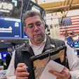 Trader at NYSE trading floor wearing headset and holding tablet with monitors displaying market data and American flag in background