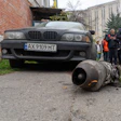 Damaged BMW sedan with detached engine component displayed on ground during investigation with officials and onlookers in urban courtyard