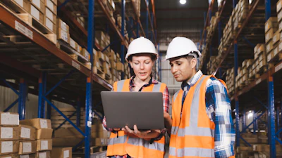 Two warehouse workers in safety gear reviewing inventory data on laptop in storage facility