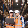 Two warehouse workers in safety gear reviewing inventory data on laptop in storage facility
