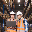 Two warehouse workers in safety gear reviewing inventory data on laptop in storage facility
