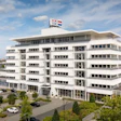 Modern white multi-story commercial building with glass windows and flags on rooftop, surrounded by landscaping and parked vehicles
