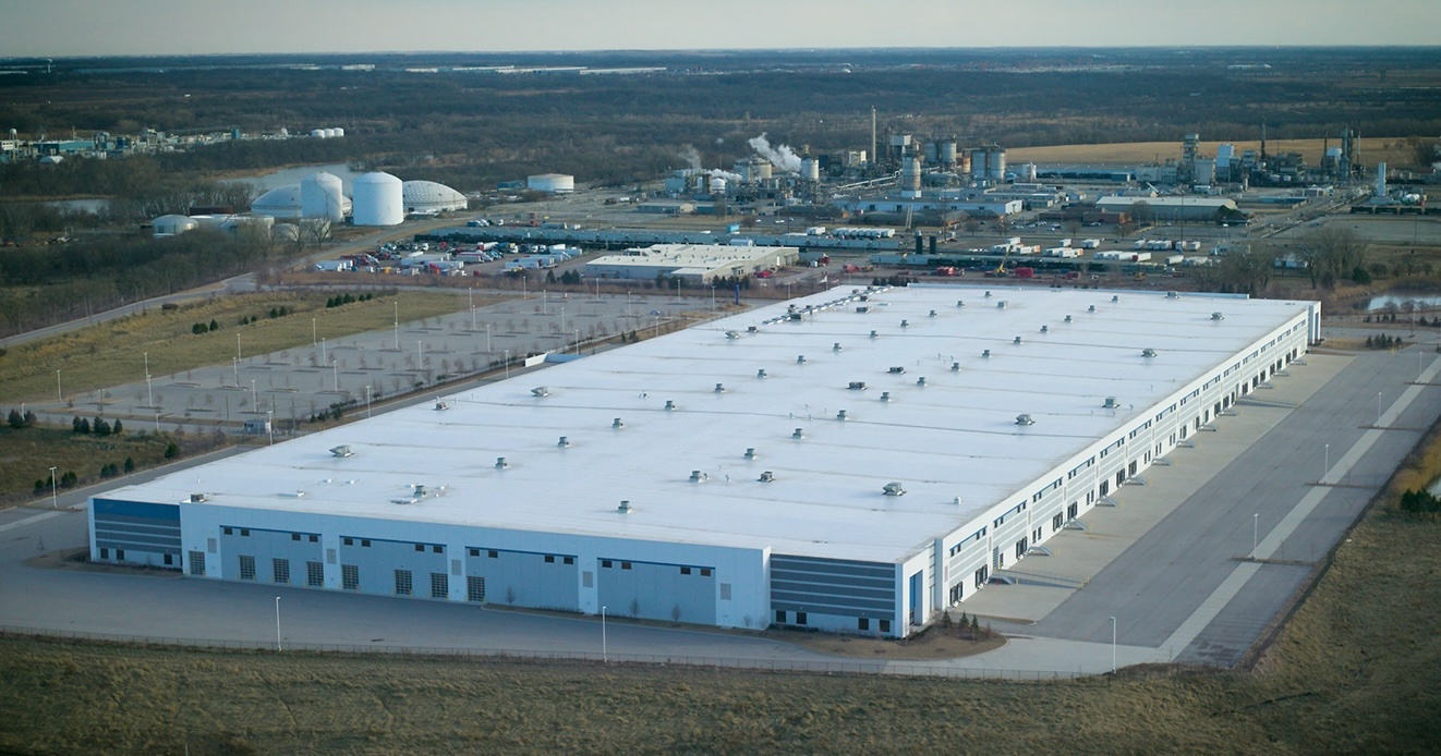 Aerial view of large industrial warehouse facility with white roof and blue walls in rural landscape